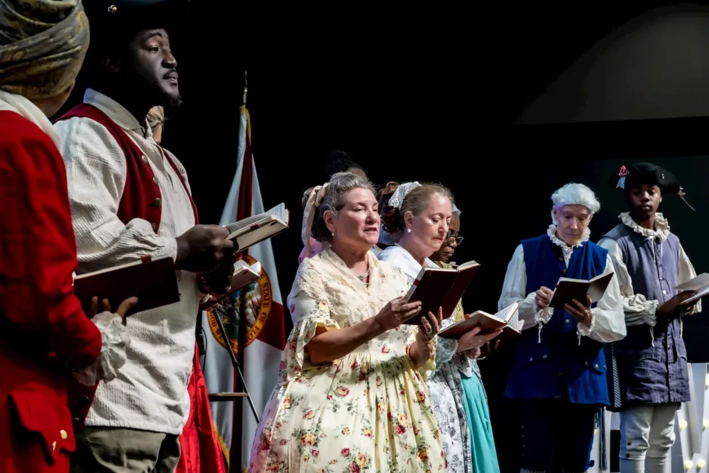 Conozca la Declaración de Independencia 1 A group of people in historical costumes stand singing or reciting from books on stage, with a flag and dark background behind them.