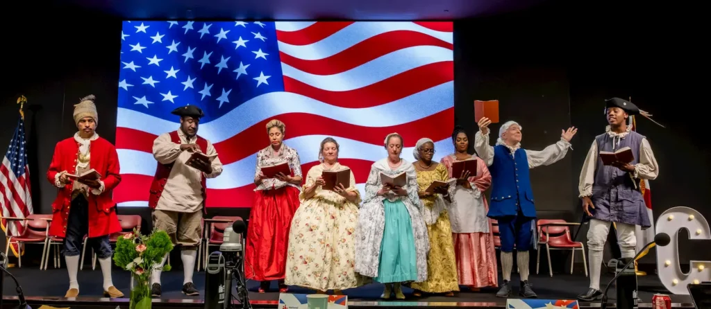 Conozca la Declaración de Independencia 2 Nine people in colonial costumes sing or read from books on stage, with a large American flag displayed behind them. A U.S. flag and chairs are visible on each side of the stage.