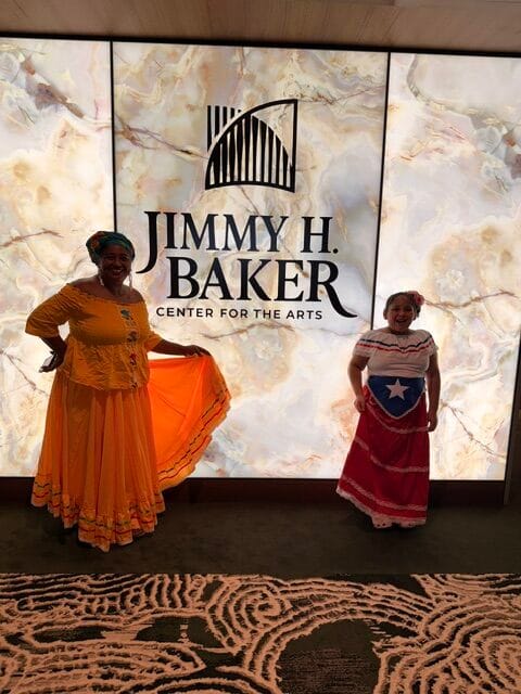 Two people in colorful traditional dresses smile and pose in front of a wall sign that reads "Jimmy H. Baker Center for the Arts." The background features a marble-like pattern.