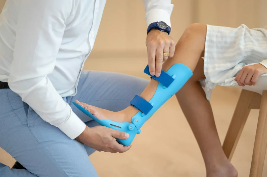 A person fits a blue orthopedic leg brace onto a child's lower leg and foot, securing it with blue straps—providing support after an ankle sprain. The child is seated, wearing a light, striped garment.
